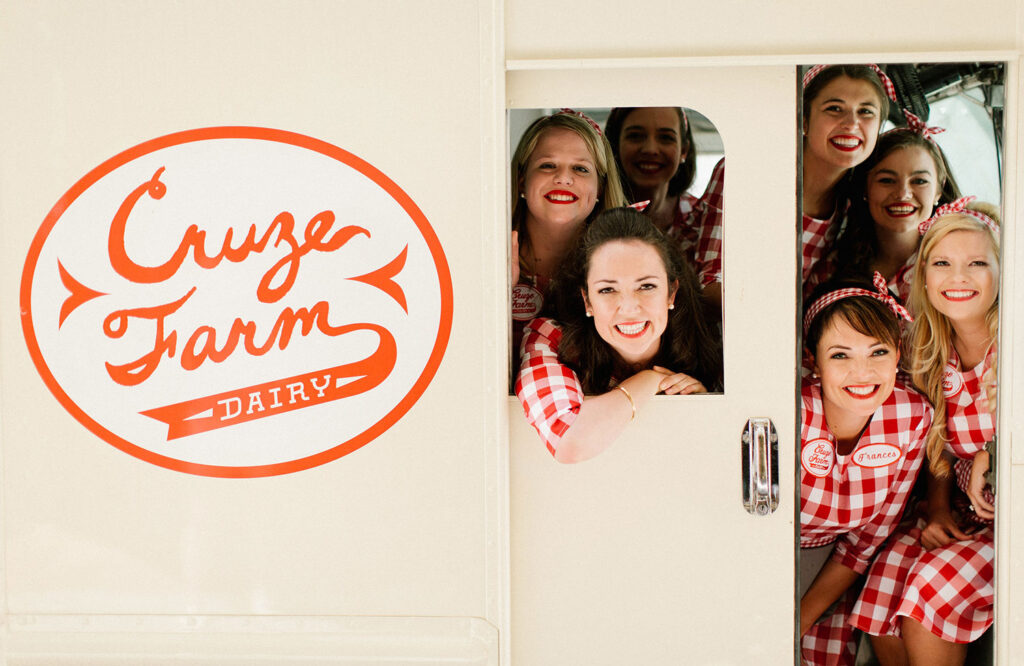 Women wearing red and white gingham dresses and headbands pose inside a cream-colored Cruze Farm Dairy truck.
