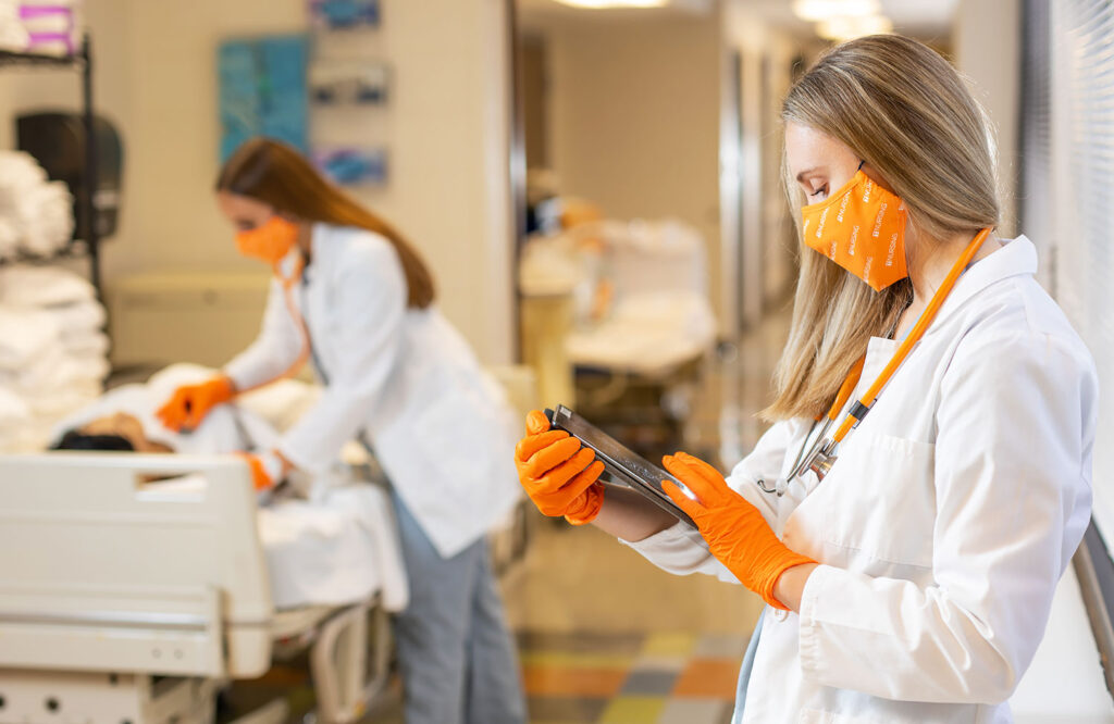 Nursing student wearing white coat, orange face covering and gloves, looks at the V-Visit Sim app on her tablet.
