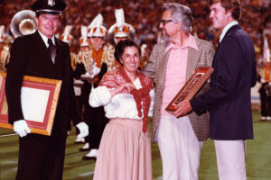 Three men and a woman stand on a football field in front of the marching band