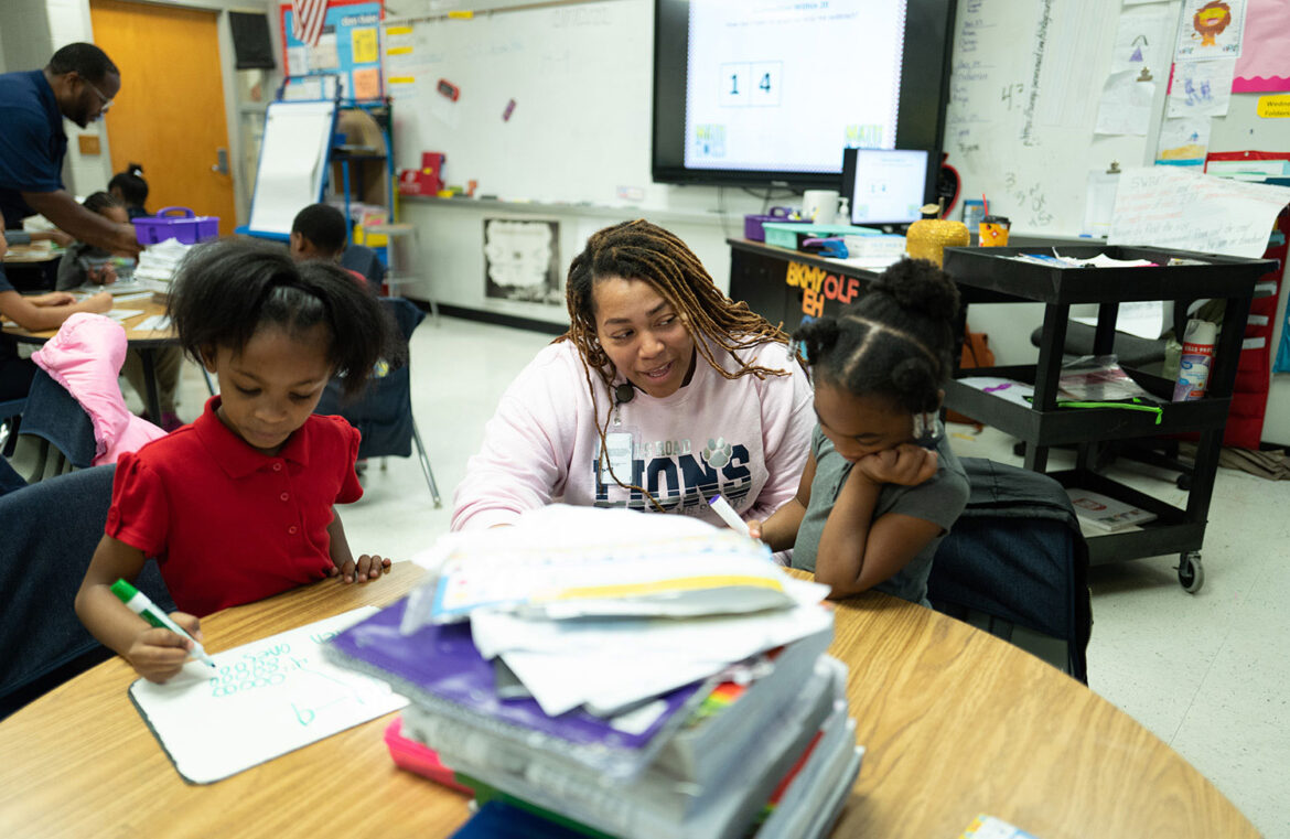 A teacher sits between two elementary school students, working at a round table