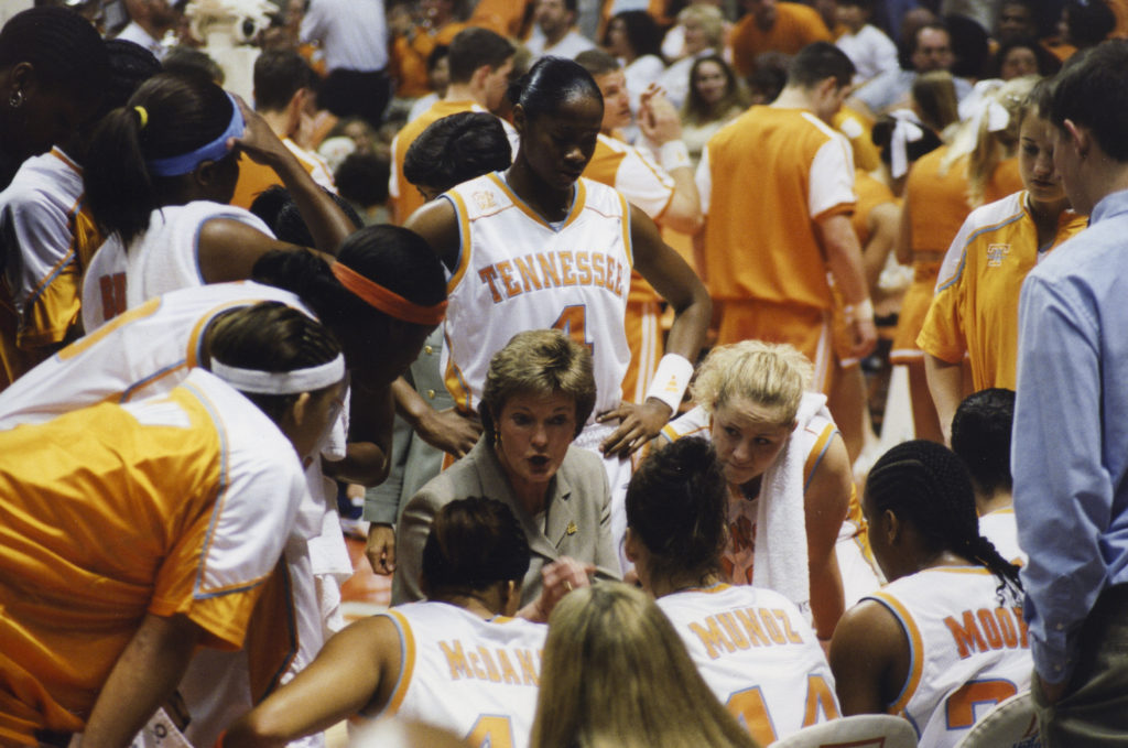 Coach Pat Summitt sits in the middle of a team of female basketball players dressed in white uniforms during a game.
