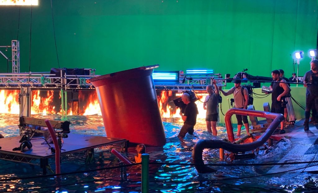A crew of people film in a pool of water on a movie set with fire behind it.