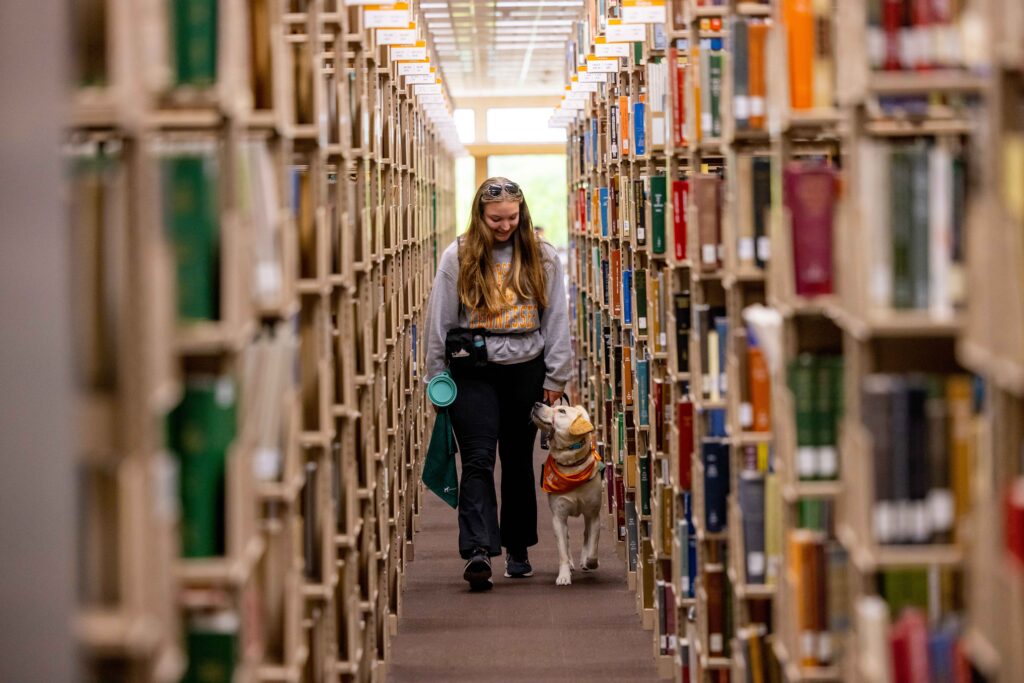 A woman walks through the library and rows of book shelves with a service dog.
