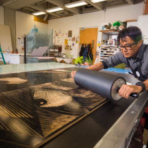 An Asian man with dark hair, wearing black-framed glasses and a black printmaking apron, pulls a large linoleum roller over a woodcut.