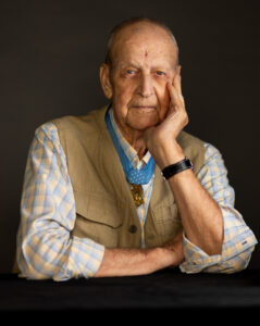 An older man sits in front of a gray background wearing a medal of honor