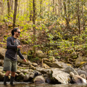 A man in a long sleeve gray shirt, wearing sunglasses, with his long brown hair pulled back by a head wrap, stands in a mountain stream fly fishing