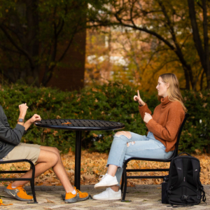 Callie Canfield and a student sit outside on an autumn day chatting in ASL.