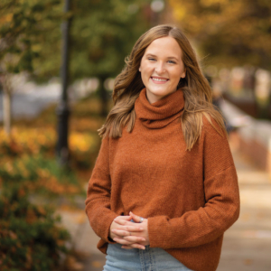 Callie Canfield outside on an autumn day, smiling, wearing an orange-brown turtleneck sweater