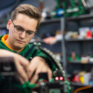 Grant Kobes, wearing glasses, an orange polo and a green jacket, concentrates on repairing a competition robot.