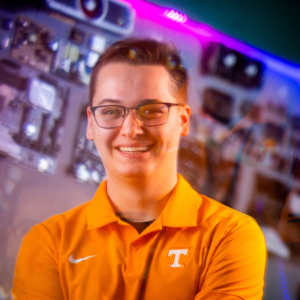 A portrait of Grant Kobes, a young white man wearing an orange polo t-shirt and glasses. 