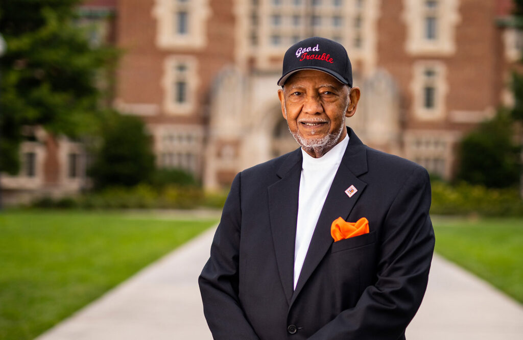 Theotis Robinson, an older Black gentleman wearing a suit and baseball hat with the words Good Trouble stitched on the front, stands in front of Ayres Hall
