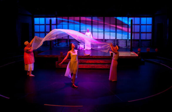 An actor walks beneath a gauzy piece of fabric being held aloft by two other actors on stage lit with purple, pink, and blue