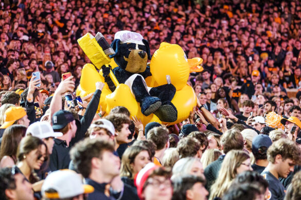Mascot Smokey, wearing a shower cap and holding a large brush, rides a giant yellow rubber ducky through the crowd of fans