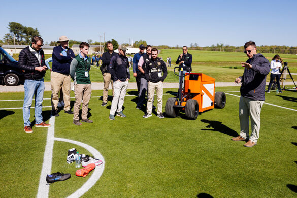 Kyley Dickson discusses the operation of the fLEX machine with a group of men—the men and machine are standing outside on an athletic field with a small pile of athletic shoes in the foreground
