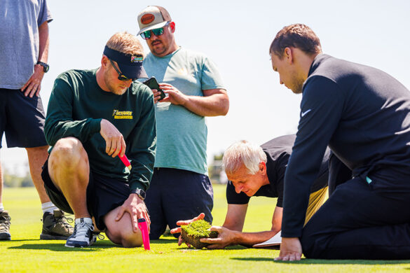John Sorochan kneels on the turfgrass surrounded by a group of men, closely examining a plug of turfgrass that he is holding in his hands.