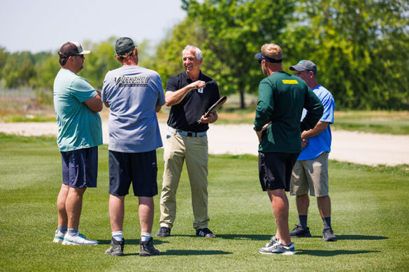 John Sorochan, standing among a group of men on bright green turfgrass and holding a clipboard, smiles and gestures with a pen