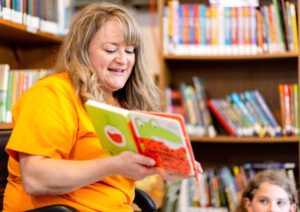 An elementary teacher wearing a bright orange shirt holds a book as she reads to elementary students
