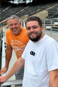 Two men smile and pose for a photo, the one on the left with gray hair and slight beard wears an orange Tennessee shirt, while the one on the right with brown hair and beard wears a white t-shirt