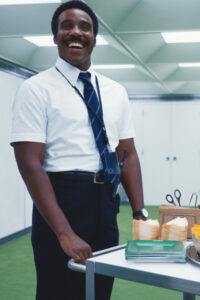 A tall Black man with a mustache wearing business casual, smiling and standing in an office environment