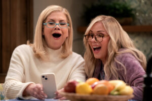 Two blonde women in glasses laugh while looking at an iPhone together
